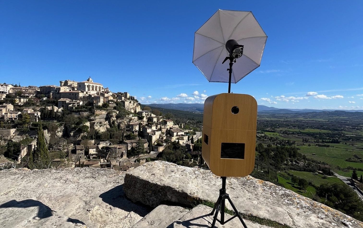 La borne photobooth Provence Photobooth avec son flash parapluie, devant un village perché du Luberon